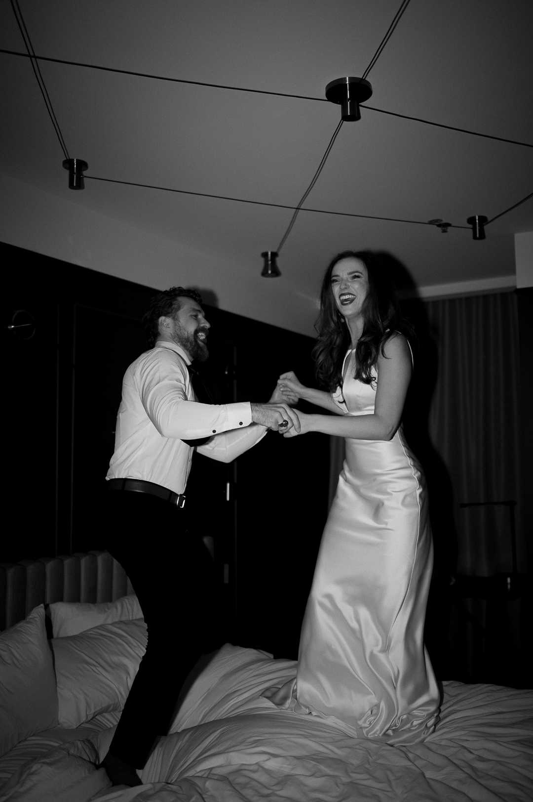 a black and white film photo with flash of a couple jumping on a hotel bad after getting married